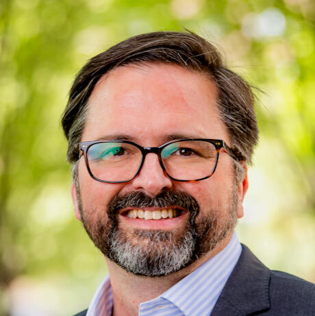 A man with dark hair and a beard is smiling at the camera. He is wearing black-framed glasses, a light blue collared shirt, and a dark jacket. The background is blurred greenery, and the image is framed in a circular shape.